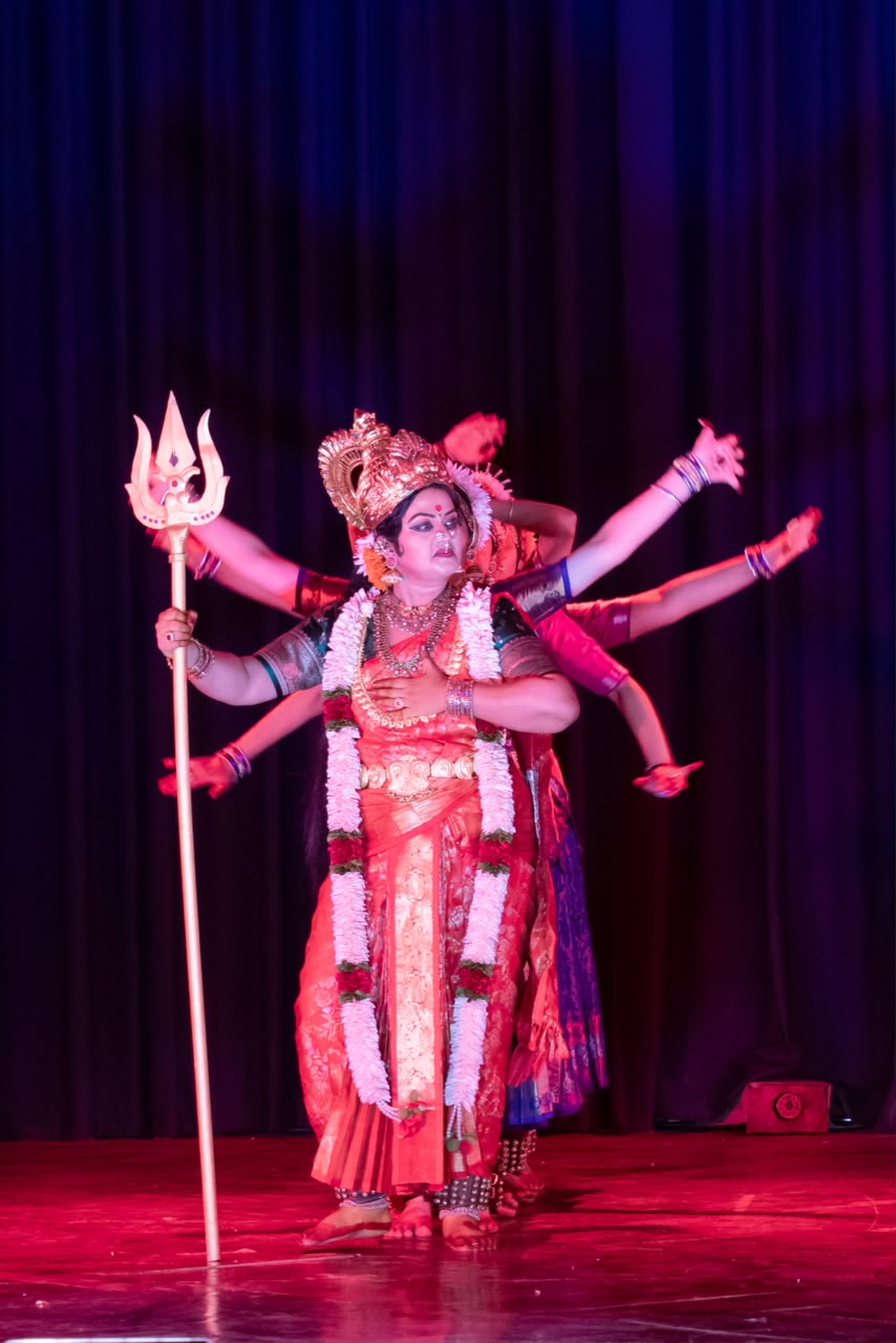 Solo dancer portraying a multi-armed goddess with a trishul, under red stage light