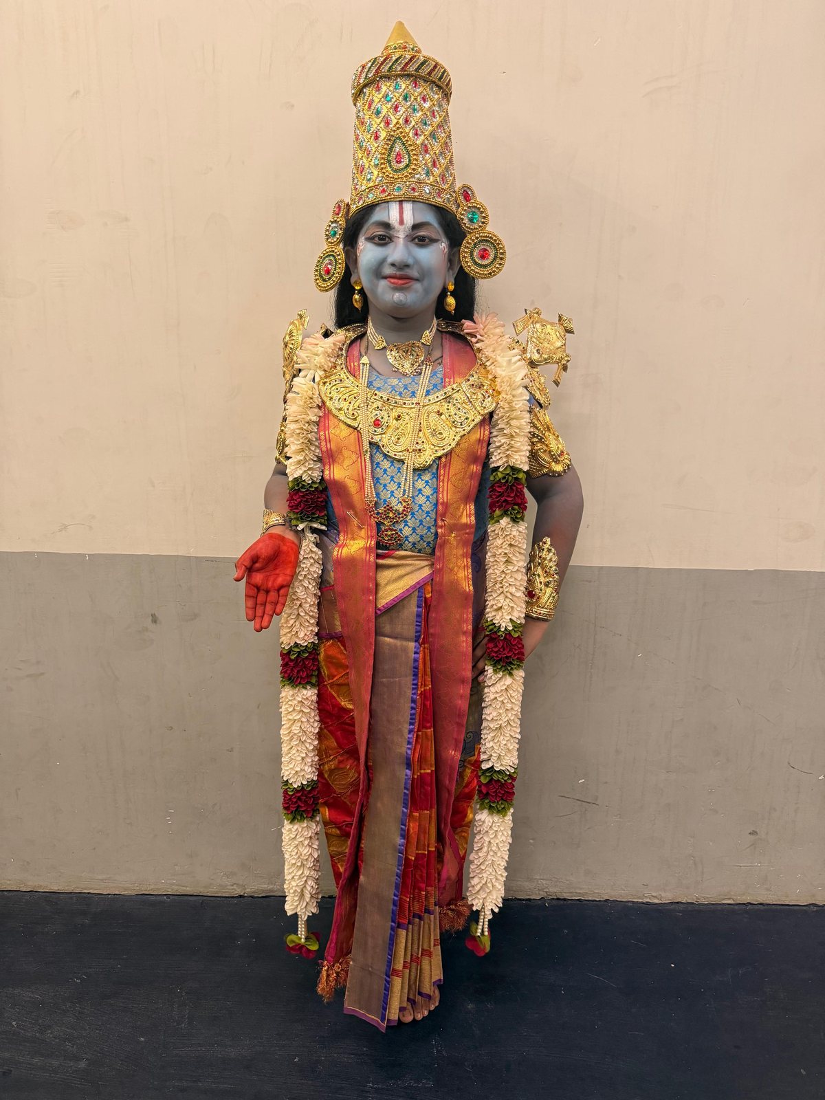 A young performer dressed as a deity, in blue makeup, gold crown, and floral garlands