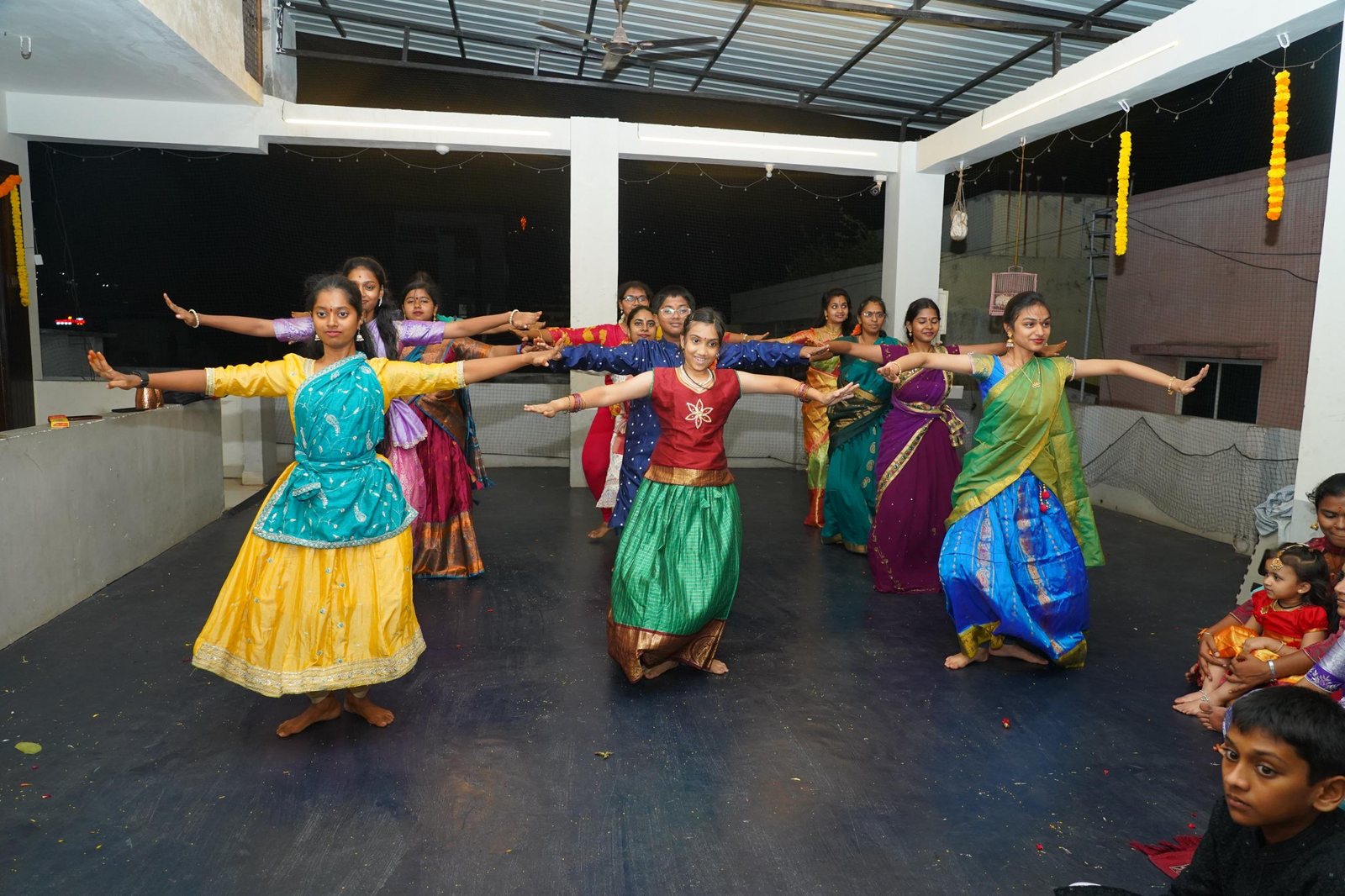 A large ensemble of dancers in colourful costumes, arms outstretched in unison on a festive rooftop stage