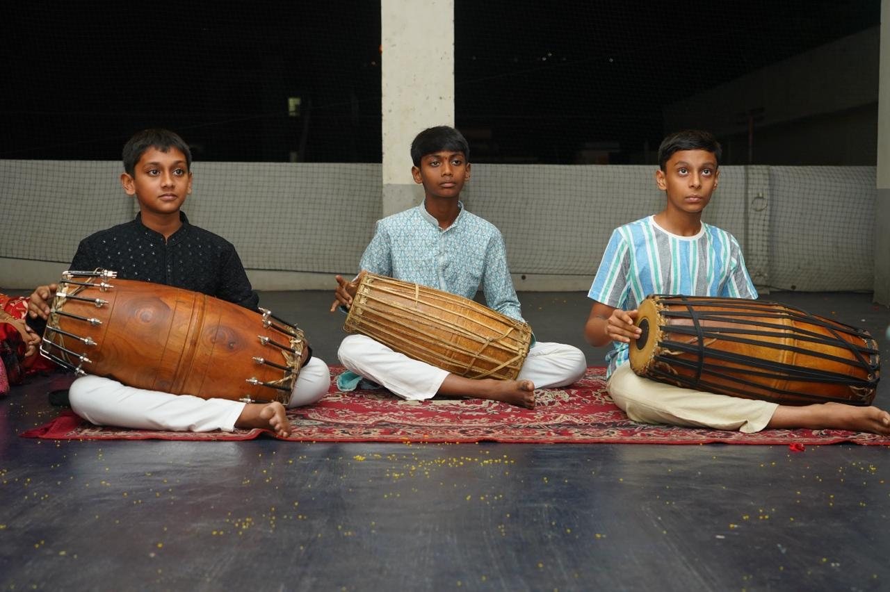 Three young mridangam students seated with their instruments on a red carpet